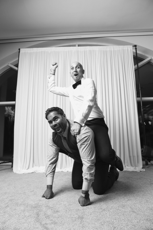 A man in a bow tie rides piggyback on a friend crawling on all fours, fist in the air