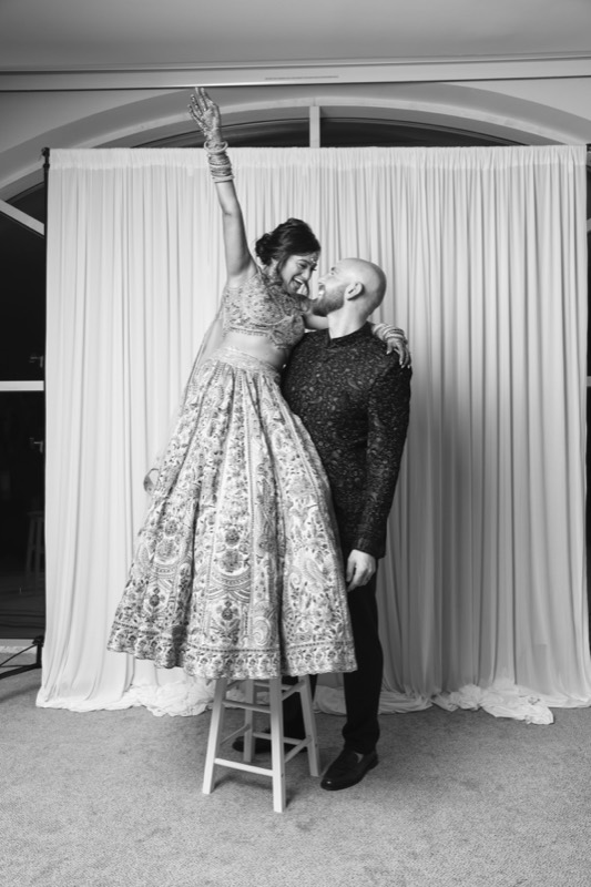 A bride in a lehenga stands on a stool, arm raised, as her groom holds her